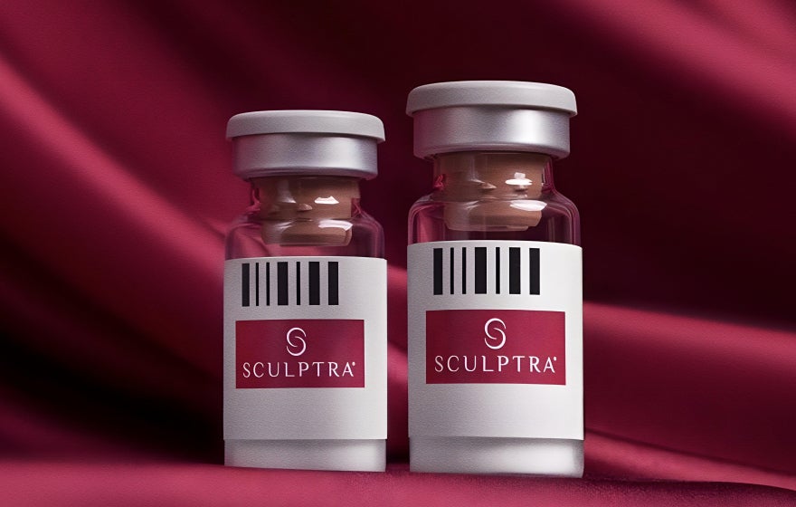 Duas ampolas transparentes do bioestimulador de colágeno Sculptra, com rótulos brancos e detalhe em vermelho que exibem o nome da marca, dispostas lado a lado sobre um elegante tecido de seda na cor vinho. A imagem foca no produto Sculptra, conhecido por estimular a produção de colágeno e elastina, essenciais para a firmeza e elasticidade da pele.