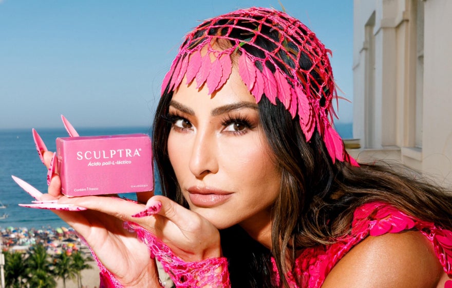 Imagem de Sabrina Sato, em um ambiente externo ensolarado com vista para o mar e palmeiras ao fundo. Ela está vestindo uma roupa vibrante em tons de rosa com detalhes em lantejoulas e acessórios que remetem a um estilo tropical e festivo. Sabrina segura uma caixa do produto Sculptra® próxima ao rosto, destacando o nome do bioestimulador de colágeno. 