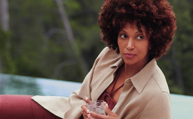 Uma mulher negra de cabelos volumosos e cacheados, usando uma camisa bege e calça vinho, relaxa em um sofá claro ao ar livre perto de uma piscina, com uma bebida em um copo transparente nas mãos. Ela olha para o lado, com uma expressão serena e confiante, resultado do uso do bioestimulador de colágeno Sculptra. Ao fundo, uma área verde desfocada