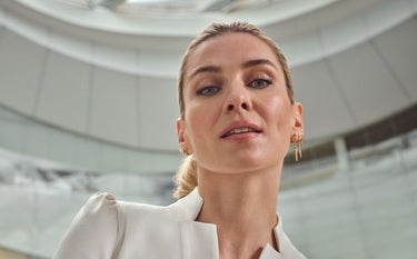 Uma mulher com pele com viço, realçada pelo uso de Restylane® Shaype™ com ácido hialurônico, é o foco desta imagem. Ela usa um vestido de lantejoulas que complementa sua aparência sofisticada. Seu olhar direto para a câmera transmite confiança, enquanto a iluminação suave e o fundo dourado contribuem para uma atmosfera de elegância e luxo. A pele lisa e firme sugere os efeitos do tratamento estético.