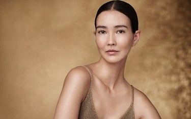 Uma mulher asiática de pele clara, com cabelo escuro preso para trás em um coque elegante, está posicionada no centro da imagem, olhando diretamente para a câmera com uma expressão serena. Ela usa uma blusa de alças finas em um tom dourado metálico, que complementa o fundo em tons de bronze e dourado, com uma textura que lembra um tecido amassado ou uma superfície irregular. A iluminação suave realça a pele da mulher.