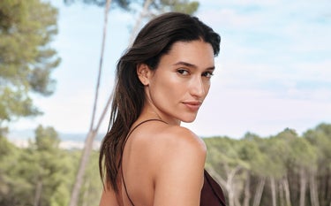 Uma mulher morena de cabelos lisos, usando uma blusa de alças finas marrom. Ao fundo, uma área verde desfocada