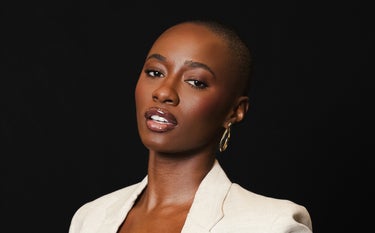 Uma mulher de pele escura e cabelo raspado, com um corte de cabelo bem curto, posa de frente para a câmera contra um fundo preto. Ela está usando um blazer de cor clara, possivelmente bege ou creme, e um brinco dourado em formato de onda na orelha direita. Seus lábios, que parecem ter sido realçados por preenchimento labial, estão levemente abertos, revelando os dentes, e têm um brilho labial que reflete a luz. A maquiagem realça seus olhos e maçãs do rosto, dando-lhe uma aparência elegante e confiante.