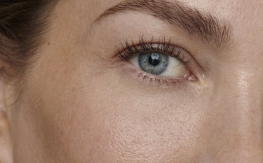 Uma imagem em close-up do rosto de uma pessoa, focando no olho esquerdo. A pele tem um tom claro e é visível a textura com alguns poros e pequenas sardas ou manchas claras. O olho, de íris azul-acinzentada, está aberto e centralizado na imagem, com a pupila escura refletindo uma fonte de luz. Os cílios superiores são longos e escuros, com um leve volume, e os cílios inferiores são mais curtos. A sobrancelha é bem definida e de cor castanha. A área da bochecha e parte da orelha são visíveis no lado esquerdo`